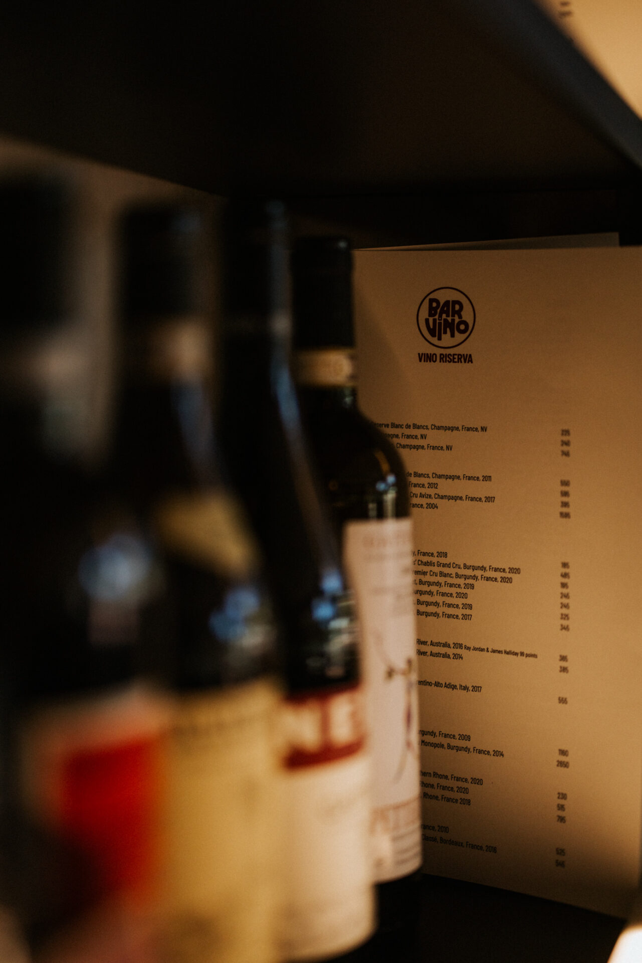 An artistic shot looking across 4 bottles off red wine out of focus towards a Bar Vino menu.