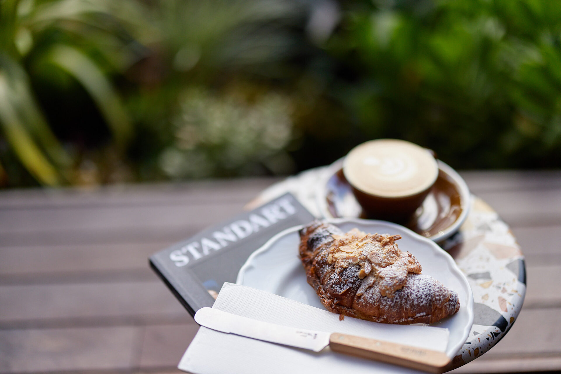 Almond Croissant on small white plate, with coffee and magazine out of focus behind.