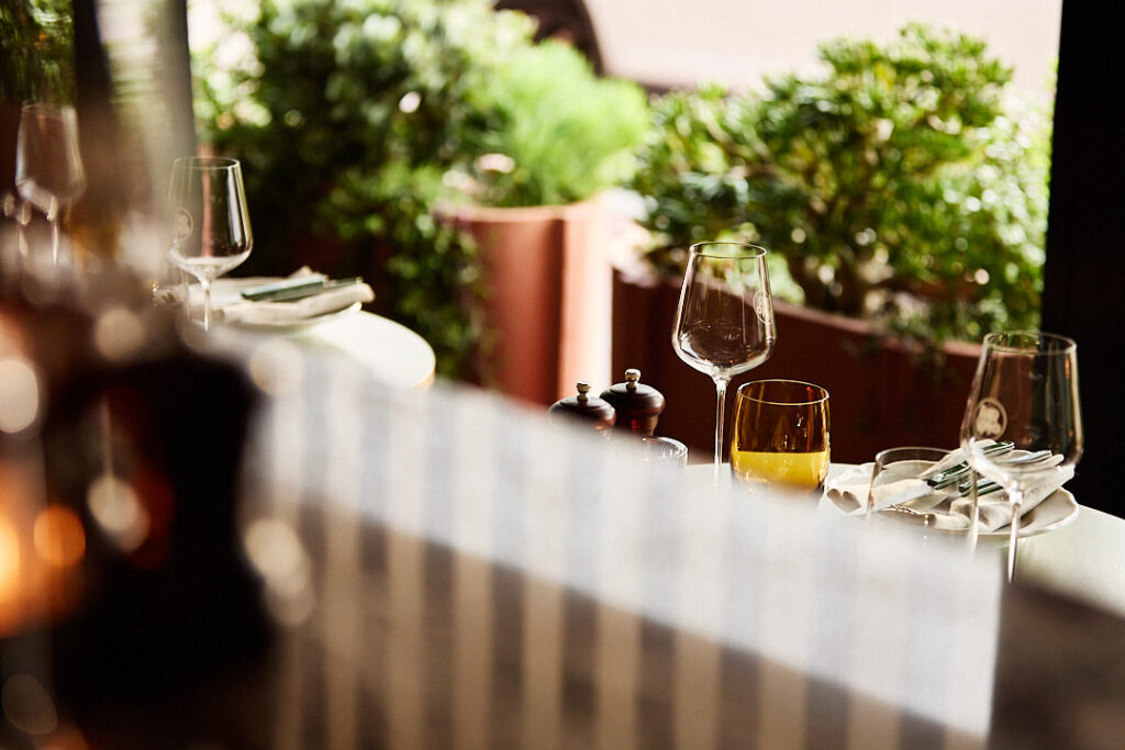 View through a window to outside to an empty chair and table with wine glasses.